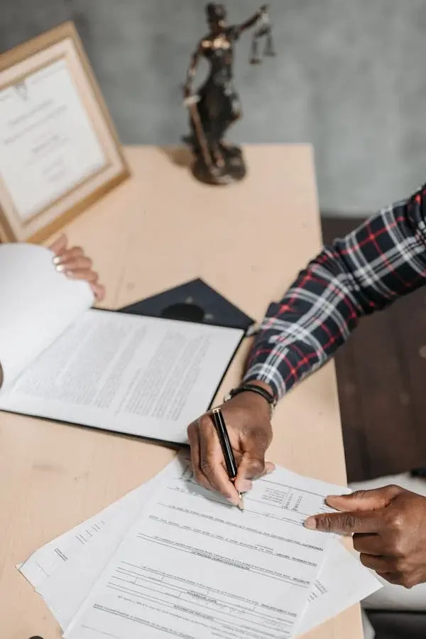 Lawyer signing intellectual property documents at office desk with legal scale and certificate in background